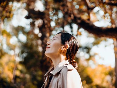 Portrait of young Asian woman having a walk in the park, enjoying the warmth of sunlight on a beautiful Autumn day outdoors and breathing fresh air with eyes closed. Relaxing in the nature under maple trees