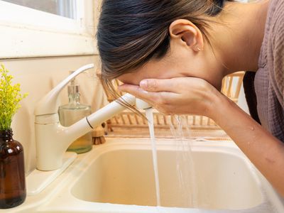 woman leans over white sink to wash face with water and natural cleanser