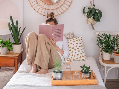 woman reads book in bed surrounded by various plants on chairs and trays