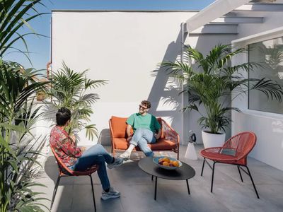 An outdoor balcony with potted plants and two people lounging and visiting