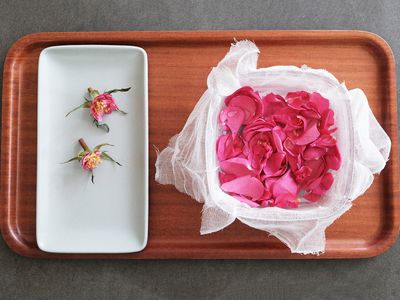 freshly picked pink rose petals in cheesecloth-lined bowl next to white tray with empty buds