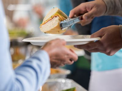 Someone holds a paper plate and uses tongs to put a subway sandwich on it, while someone else holds their hand out for the plate