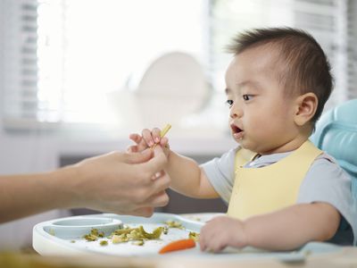 Asian baby grabs veggies from adult hand while sitting in high chair