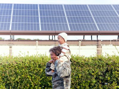 A man carries a little boy on his shoulders, both laugh happily. Solar panels and plants can be seen in the background.