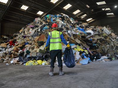 worker in construction vest holds bag of trash in front of pile of plastic rubbish