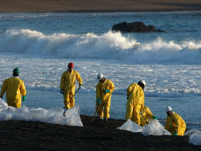 Oil spill cleanup workers in yellow hazmat gear and hard hats shovel oil-contaminated sand along a beach with clear plastic bags in foreground.