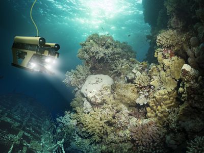 A robot exploring an underwater reef with headlights. A cord hangs from the robot.