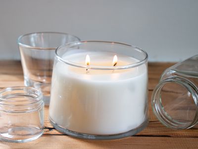 lit white candle on wooden table surrounded by empty glass candle jars