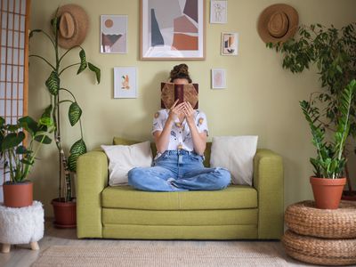 woman reads book on green couch surrounded by various large house plants