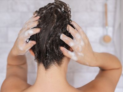 back of woman in white tiled shower with hands scrubbing shampoo in hair