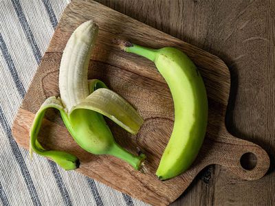 two green bananas on wooden cutting board