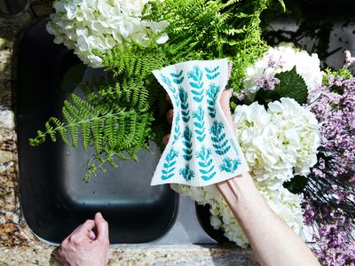 A hand holds a reusable dishcloth over a sink filled with flowers