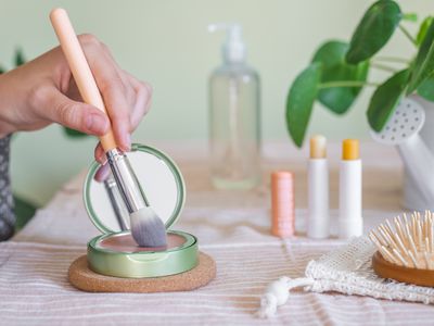 woman rubs makeup brush in green makeup blush container next to beauty products