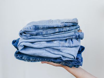 hand holds out multiple pairs of denim jeans stacked on top of each other in front of blank wall